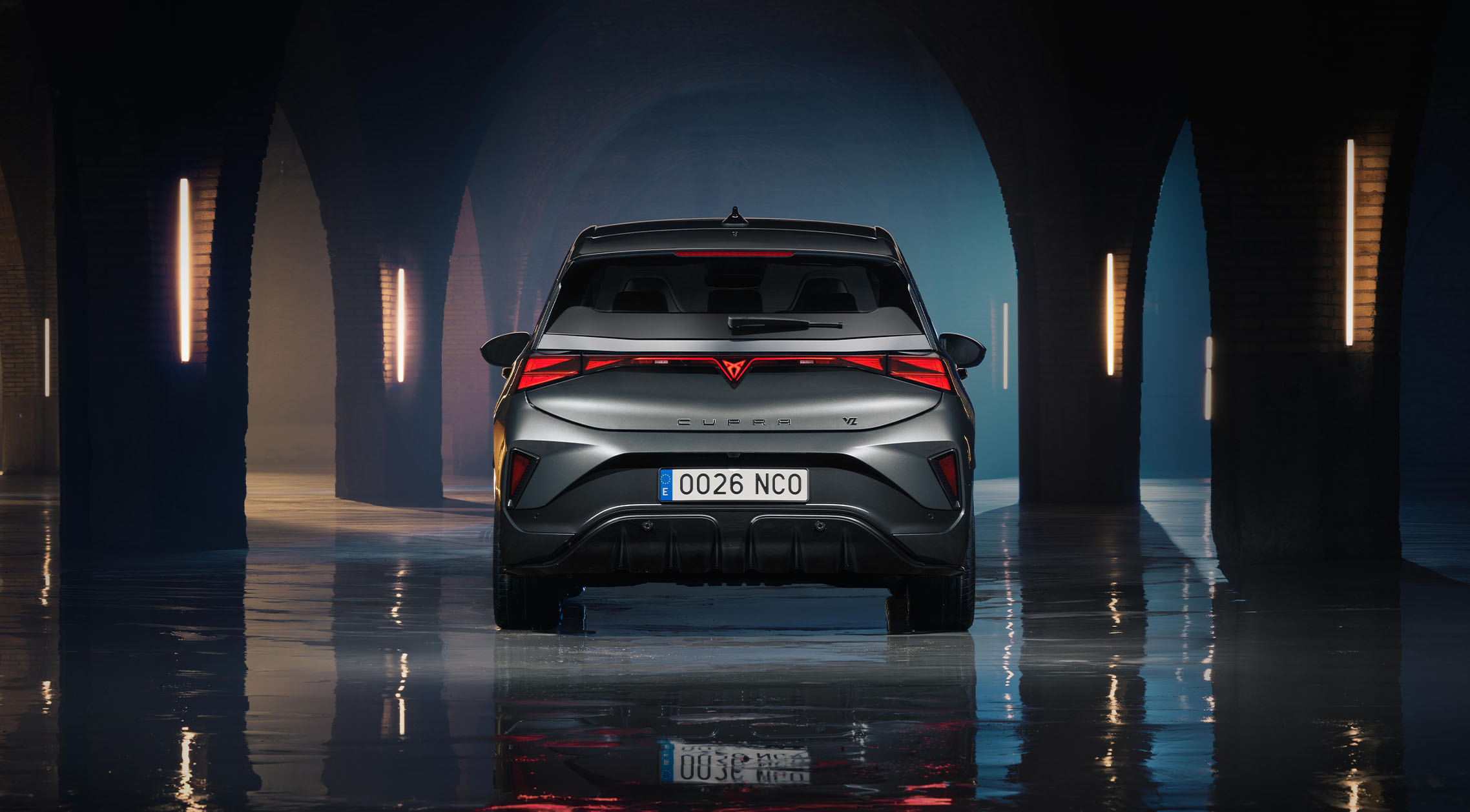Vista posteriore diretta di una CUPRA Born VZ in colore Timanfaya Grey, parcheggiata in uno spazio industriale scuro e con volte ad arco in pietra, illuminato da barre luminose al neon verticali. L’immagine mette in evidenza la striscia luminosa LED posteriore continua, il logo distintivo CUPRA e il riflesso del veicolo su un pavimento in cemento lucido e bagnato.