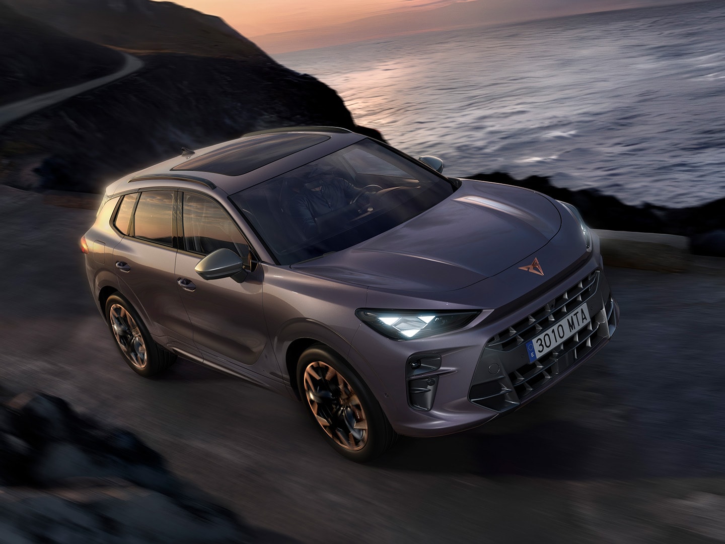 CUPRA Terramar, con Dynamic Chassis Control (DCC), che guida su una strada costiera con vernice Dark Void e leghe di Hadron Copper da 20". 